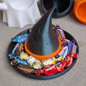 May include: A black witch's hat with moon and star details sits on an orange ring on a black tray filled with wrapped sweets. Halloween-themed candy dish with ghost, bat, and pumpkin-shaped containers in the background.