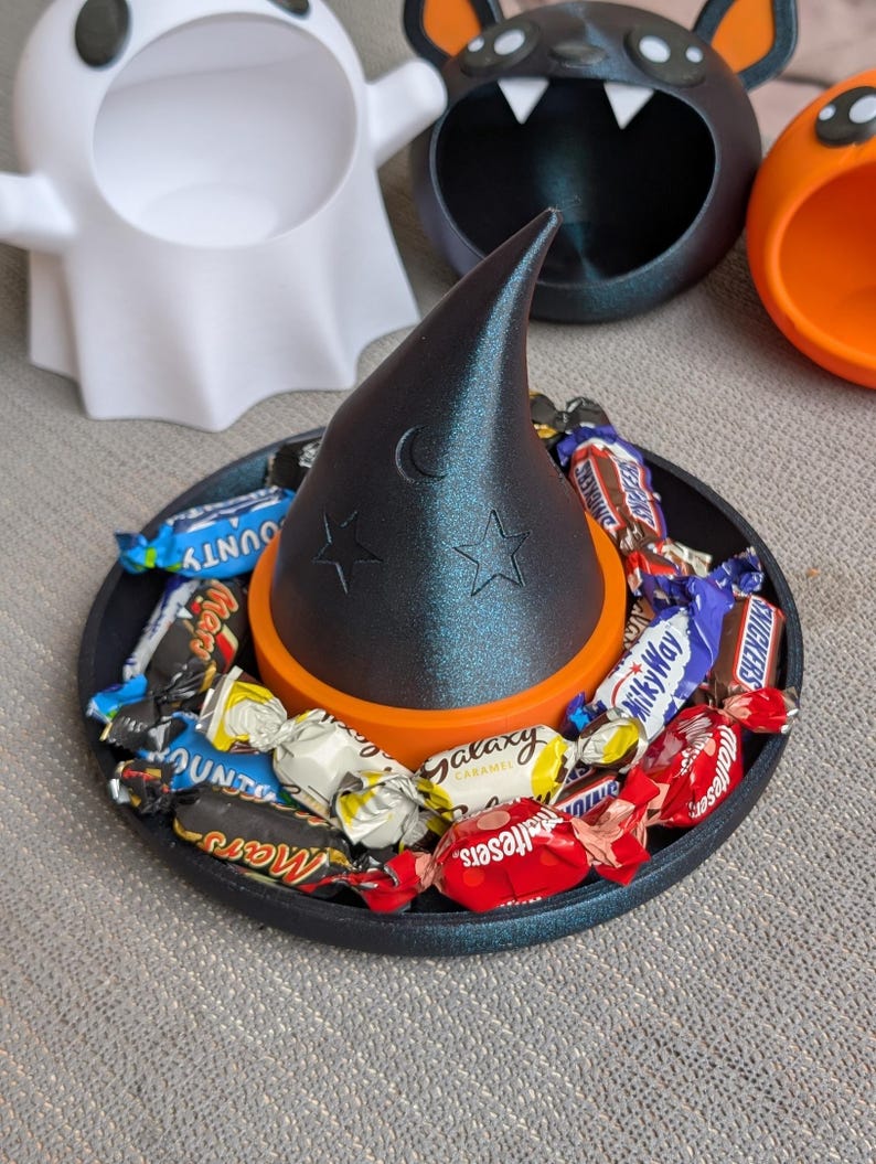 May include: A black witch's hat with moon and star details sits on an orange ring on a black tray filled with wrapped sweets. Halloween-themed candy dish with ghost, bat, and pumpkin-shaped containers in the background.