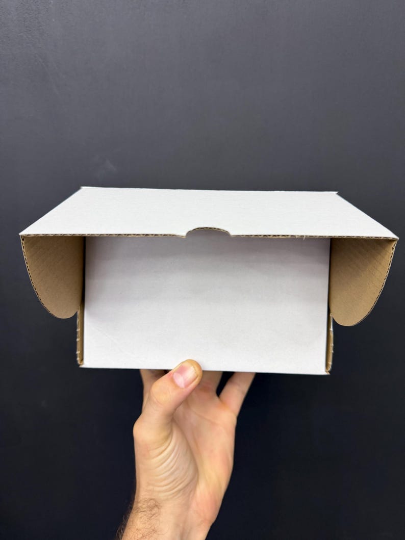 May include: A white cardboard box with the flaps open, held by a hand. The interior of the box is white, while the inside of the flaps is brown. The background is a solid black.
