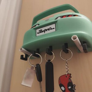 May include: A mint green car-shaped key holder with the word "Supra" on the license plate. The key holder has three key hooks and two exhaust pipe-shaped holders. Several keys and keychains hang from the hooks.