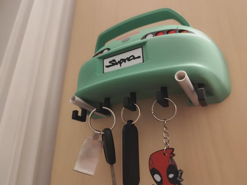 May include: A mint green car-shaped key holder with the word "Supra" on the license plate. The key holder has three key hooks and two exhaust pipe-shaped holders. Several keys and keychains hang from the hooks.