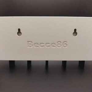 May include: An off-white rectangular key holder for wall mounting, featuring four black pegs. It has two keyhole slots and the engraved text "Becce86". The key holder is designed for keys.