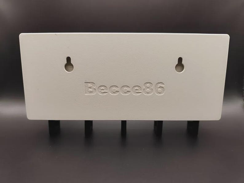 May include: An off-white rectangular key holder for wall mounting, featuring four black pegs. It has two keyhole slots and the engraved text "Becce86". The key holder is designed for keys.