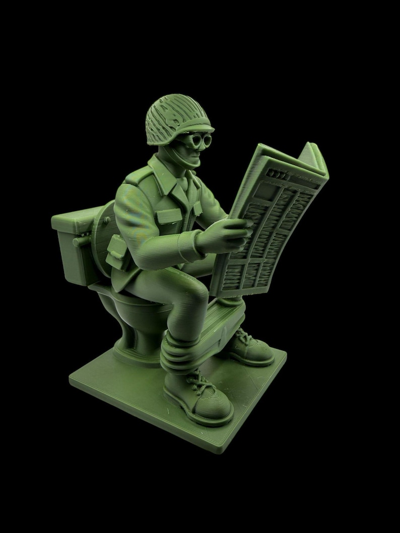 May include: A green figurine of a soldier sitting on a toilet, reading a newspaper. The soldier wears a helmet, glasses, and military uniform. The figurine is on a square base, with a black background.