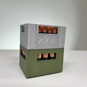 May include: Two stacked battery holders, one grey and one olive green, each filled with batteries. The grey holder is labelled "AAA", and the green holder is labelled "AA". The batteries are copper-coloured.