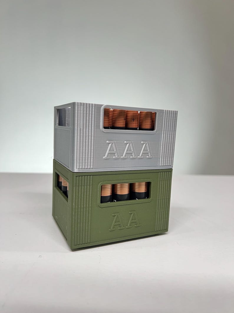 May include: Two stacked battery holders, one grey and one olive green, each filled with batteries. The grey holder is labelled "AAA", and the green holder is labelled "AA". The batteries are copper-coloured.