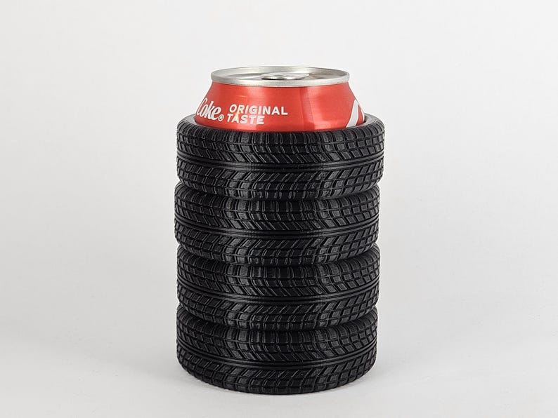 May include: A black tire-shaped can cooler with five stacked tires. The cooler is holding a red can of Coca-Cola with the text "Original Taste" on it.