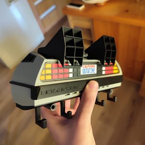 Pode incluir: Um porta-chaves cinza e preto em forma de DeLorean com a placa "OUTATIME". O porta-chaves tem aberturas pretas na parte superior e uma s&eacute;rie de luzes coloridas na parte traseira. O porta-chaves &eacute; segurado na m&atilde;o de uma pessoa.
