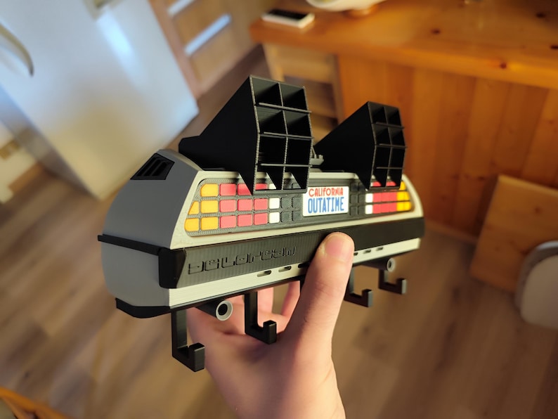 Pode incluir: Um porta-chaves cinza e preto em forma de DeLorean com a placa "OUTATIME". O porta-chaves tem aberturas pretas na parte superior e uma s&eacute;rie de luzes coloridas na parte traseira. O porta-chaves &eacute; segurado na m&atilde;o de uma pessoa.