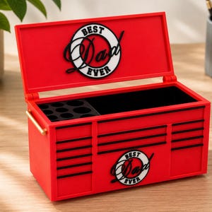 May include: A red toolbox-shaped box with the words "Best Dad Ever" in a circular design on the lid and front. The box has black accents and a black interior. It has a gold-colored handle and is open, revealing compartments.