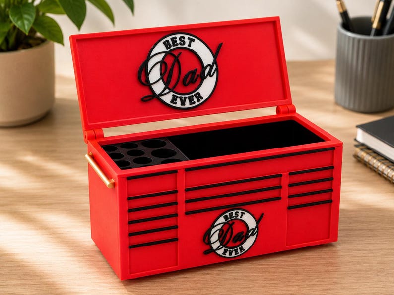 May include: A red toolbox-shaped box with the words "Best Dad Ever" in a circular design on the lid and front. The box has black accents and a black interior. It has a gold-colored handle and is open, revealing compartments.