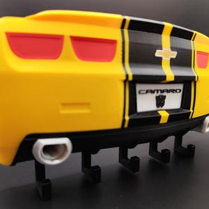 Puede incluir: Gancho de pared con forma de Camaro amarillo y negro con luces traseras rojas. El gancho tiene una franja negra en el centro y la palabra "Camaro" en la matr&iacute;cula. Tiene cuatro ganchos negros y dos tubos de escape plateados.