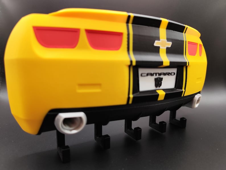 Puede incluir: Gancho de pared con forma de Camaro amarillo y negro con luces traseras rojas. El gancho tiene una franja negra en el centro y la palabra "Camaro" en la matr&iacute;cula. Tiene cuatro ganchos negros y dos tubos de escape plateados.