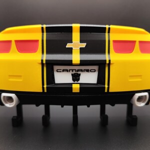 Puede incluir: Parte trasera de un coche de juguete amarillo y negro con luces traseras rojas. El coche tiene una franja negra en el centro y la palabra "CAMARO" en la matr&iacute;cula. La matr&iacute;cula tambi&eacute;n tiene un logotipo de Transformers.