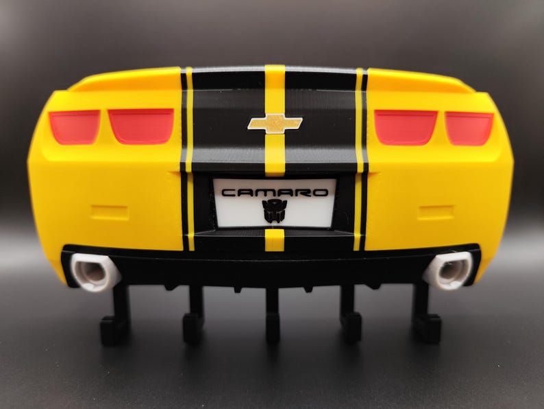 Puede incluir: Parte trasera de un coche de juguete amarillo y negro con luces traseras rojas. El coche tiene una franja negra en el centro y la palabra "CAMARO" en la matr&iacute;cula. La matr&iacute;cula tambi&eacute;n tiene un logotipo de Transformers.