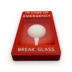 May include: A novelty item featuring a white golf ball on a tee inside a red box. The box has the words "IN CASE OF EMERGENCY" and "BREAK GLASS" in white text. The item is designed to resemble a fire alarm.