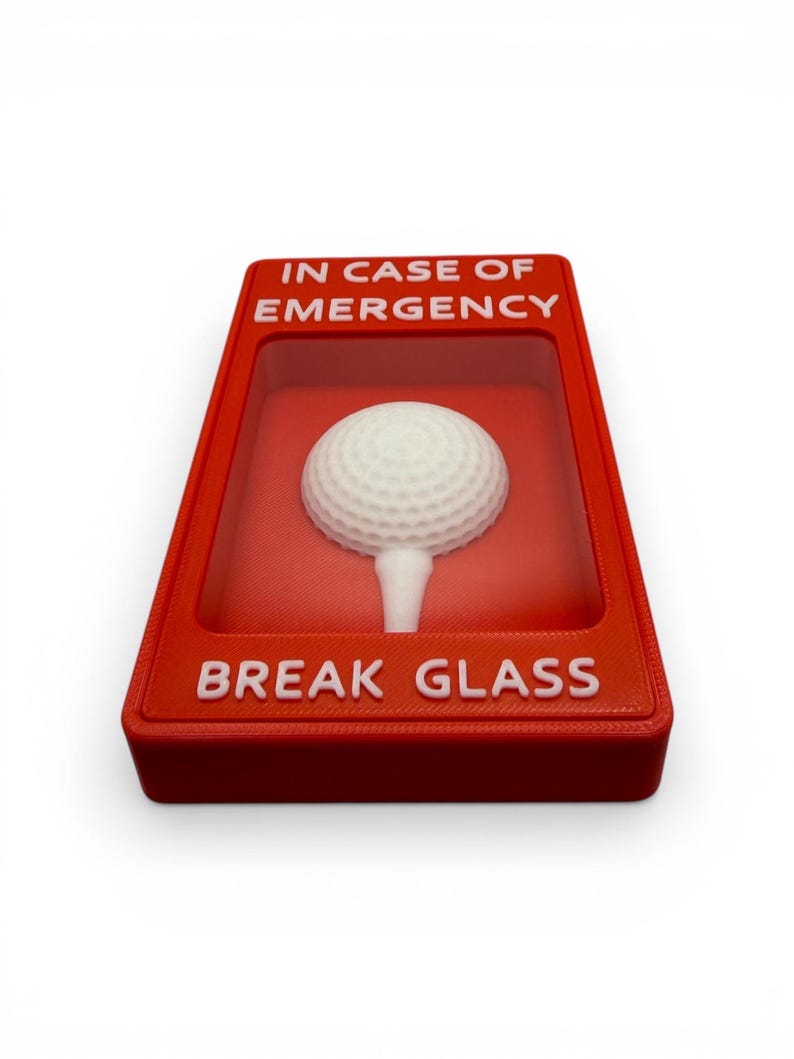 May include: A novelty item featuring a white golf ball on a tee inside a red box. The box has the words "IN CASE OF EMERGENCY" and "BREAK GLASS" in white text. The item is designed to resemble a fire alarm.