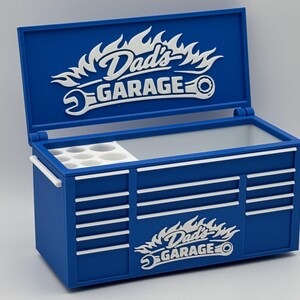 May include: A blue toolbox with white accents and the text "Dad's Garage" in a flame and wrench design. The toolbox has a hinged lid and multiple drawer-like features. It includes a white tray with circular compartments.