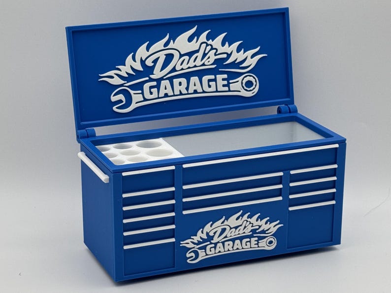 May include: A blue toolbox with white accents and the text "Dad's Garage" in a flame and wrench design. The toolbox has a hinged lid and multiple drawer-like features. It includes a white tray with circular compartments.