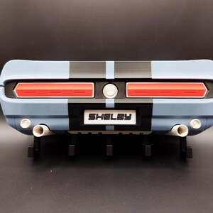 K&ouml;nnte beinhalten: Heck eines hellblauen und schwarzen Modellautos mit roten R&uuml;ckleuchten und dem Wort "Shelby" auf einem wei&szlig;en Schild. Das Modell hat doppelte Auspuffrohre und schwarze Streifen.