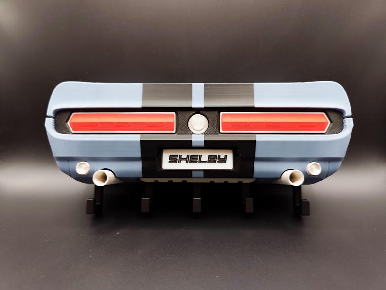 K&ouml;nnte beinhalten: Heck eines hellblauen und schwarzen Modellautos mit roten R&uuml;ckleuchten und dem Wort "Shelby" auf einem wei&szlig;en Schild. Das Modell hat doppelte Auspuffrohre und schwarze Streifen.