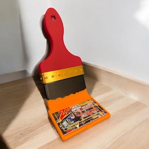 Puede incluir: Un soporte en forma de pincel rojo y negro con un porta tarjetas de visita. El porta tarjetas de visita es naranja y tiene el texto "Zut Remarkable 3D Printing & Prototyping Services" impreso en &eacute;l.