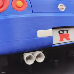 May include: Close-up of a blue toy car's rear end, featuring red taillights with orange centers. The car has a white license plate with "GT R" in red. Dual exhaust pipes are visible below the bumper.