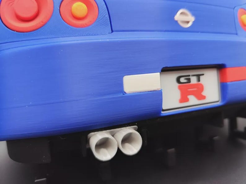 May include: Close-up of a blue toy car's rear end, featuring red taillights with orange centers. The car has a white license plate with "GT R" in red. Dual exhaust pipes are visible below the bumper.