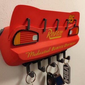 May include: Red key holder shaped like Lightning McQueen's spoiler, with the number 95 and the word "Rusteze" in yellow. It has hooks for keys and the text "Medicated Bumper Ointment".