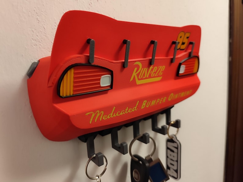 May include: Red key holder shaped like Lightning McQueen's spoiler, with the number 95 and the word "Rusteze" in yellow. It has hooks for keys and the text "Medicated Bumper Ointment".