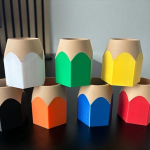 Puede incluir: Ocho porta lápices con forma de lápiz en diferentes colores: negro, blanco, verde, amarillo, naranja, azul y rojo. Los porta lápices están hechos de plástico y tienen una forma hexagonal.