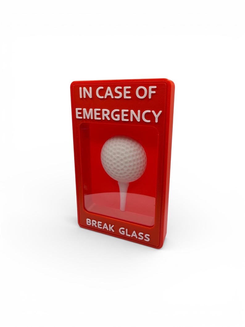 May include: A red box with the text "IN CASE OF EMERGENCY" and "BREAK GLASS" in white. Inside the box is a golf ball on a tee. The golf ball is white and has a dimpled surface.