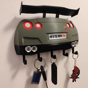Pode incluir: Um porta-chaves em forma de carro verde e preto com um spoiler, luzes traseiras e tubos de escape. O porta-chaves tem a palavra "NISMO" e inclui ganchos para pendurar chaves. Várias chaves e porta-chaves estão pendurados nos ganchos.