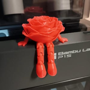 May include: A 3D printed red rose with articulated legs and arms sitting on a black 3D printer with the text "Bambu Lab P1S" on the side.