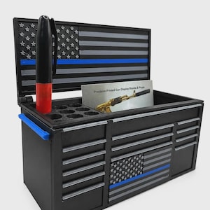 May include: A black miniature tool chest organizer featuring a thin blue line flag design on the lid and front. The unit includes a pen holder with a black and red marker, a storage compartment, and a card labeled Precision-Printed Gun Display Stands & Props.