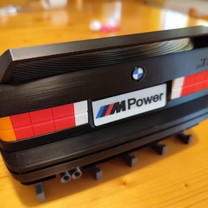 May include: Black model of a BMW M3 rear end, featuring a spoiler, taillights with red, white, and orange sections, and the "M Power" logo. The model includes exhaust pipes and the BMW emblem.