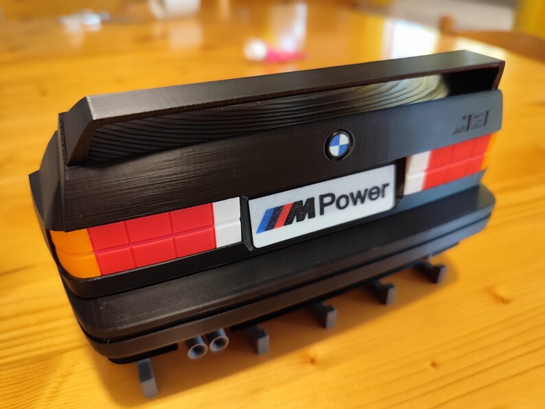 May include: Black model of a BMW M3 rear end, featuring a spoiler, taillights with red, white, and orange sections, and the "M Power" logo. The model includes exhaust pipes and the BMW emblem.