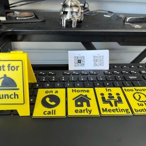 May include: A yellow sign that says "Out for Lunch" with a black graphic of a covered plate. There are also yellow signs with black graphics and text that say "on a call", "Home early", "In Meeting", and "too busy to be bothered".