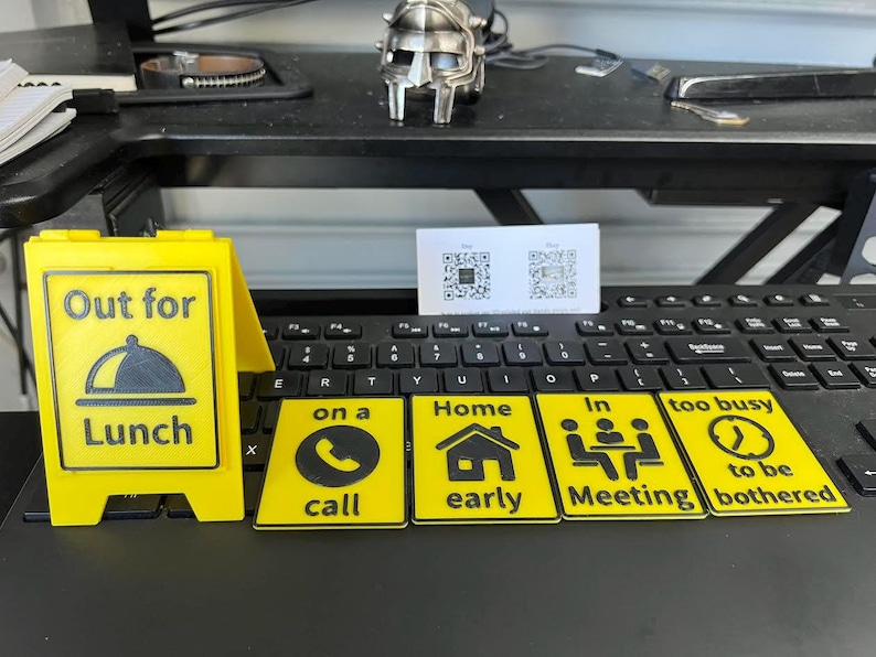 May include: A yellow sign that says "Out for Lunch" with a black graphic of a covered plate. There are also yellow signs with black graphics and text that say "on a call", "Home early", "In Meeting", and "too busy to be bothered".