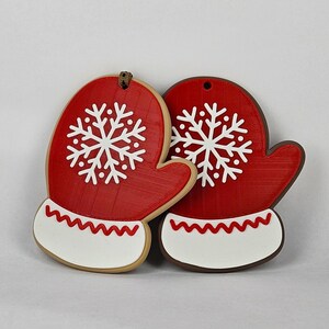 May include: Two red mitten-shaped Christmas ornaments with white snowflake designs. Each mitten has a white cuff with a red zigzag pattern. The ornaments are made of a textured material and have a hole at the top for hanging. Festive holiday decor.