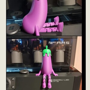 May include: A 3D-printed eggplant figurine in purple with a green stem. The figurine has articulated arms and legs. The eggplant is displayed on a black surface with a computer in the background. The text "Bambu Lab P1S" is visible.