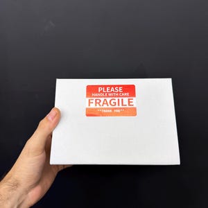 May include: A white rectangular box with a red and orange "FRAGILE" sticker. The sticker reads "PLEASE HANDLE WITH CARE" and "THANK YOU". The box is held against a black background.