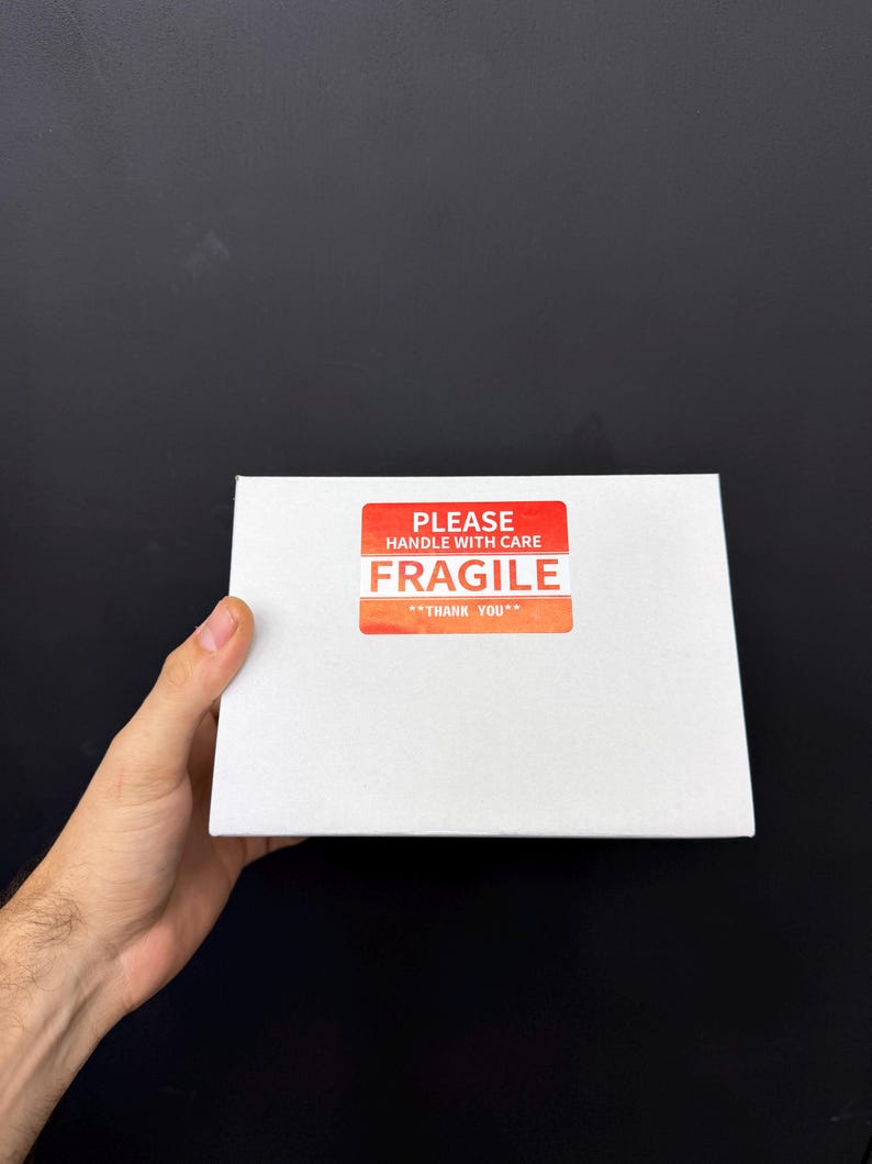 May include: A white rectangular box with a red and orange "FRAGILE" sticker. The sticker reads "PLEASE HANDLE WITH CARE" and "THANK YOU". The box is held against a black background.
