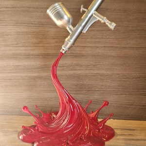 May include: A silver airbrush with a red paint splash design. The airbrush is angled, appearing to spray a stream of red paint that forms a splash. The base is a glossy red, with the airbrush details clearly visible.