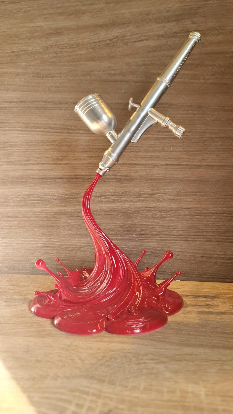 May include: A silver airbrush with a red paint splash design. The airbrush is angled, appearing to spray a stream of red paint that forms a splash. The base is a glossy red, with the airbrush details clearly visible.