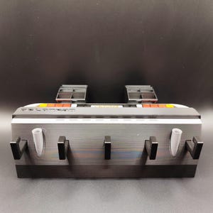 Pode incluir: Um organizador de parede preto e cinza com v&aacute;rios ganchos e dois suportes cil&iacute;ndricos. A parte superior apresenta um design branco, laranja e preto. O organizador foi projetado para guardar itens.