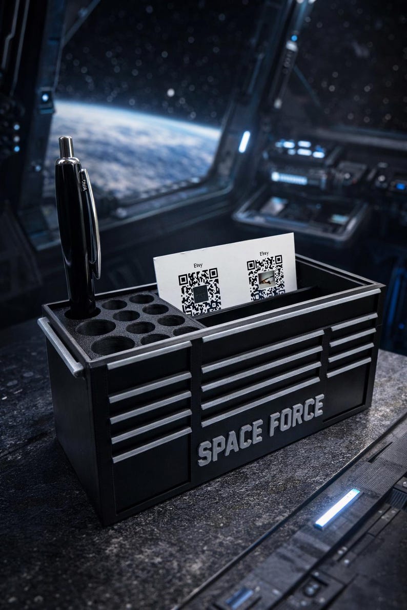 May include: Black desk organizer with the text "SPACE FORCE" in white. The organizer has a section for pens and business cards. A black pen is in the pen holder. Two cards with QR codes are in the business card holder.