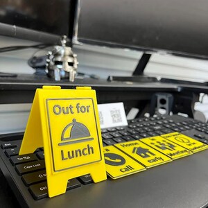 May include: A bright yellow sign with the words "Out for Lunch" and a graphic of a covered plate. The sign is propped up on a laptop keyboard. Additional yellow cards with icons and text are also on the keyboard.