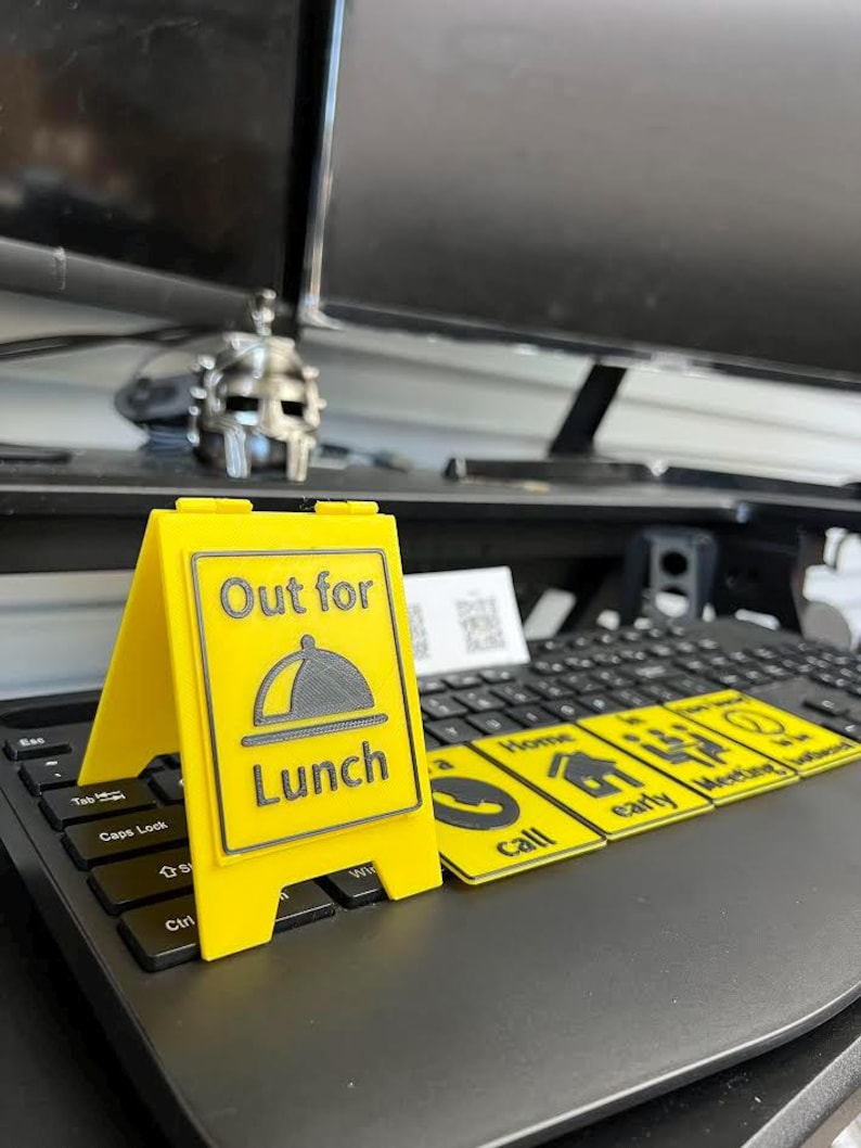 May include: A bright yellow sign with the words "Out for Lunch" and a graphic of a covered plate. The sign is propped up on a laptop keyboard. Additional yellow cards with icons and text are also on the keyboard.