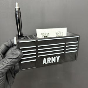 May include: Black desk organizer with the word "ARMY" in white block letters. The organizer has a slot for business cards and a hole for a pen. A black pen is in the hole. The organizer is made of a black material.